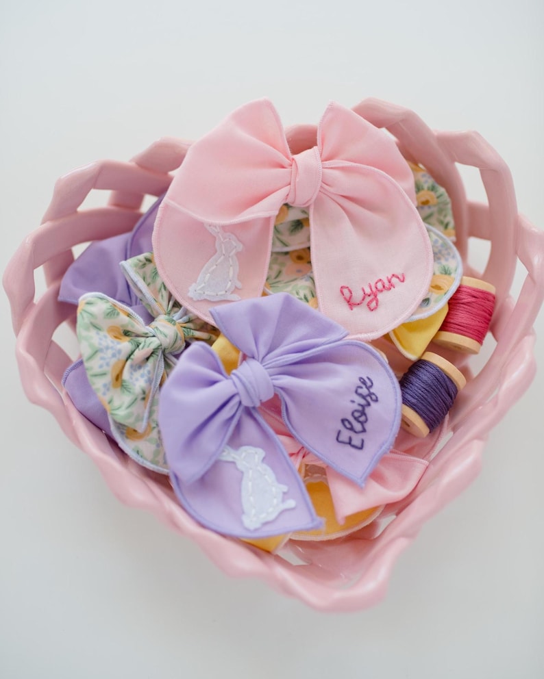 May include: A pink heart-shaped basket containing fabric bows in pink, lavender, and floral patterns. The bows feature embroidered white rabbit silhouettes and the names Ryan and Eloise stitched in thread. Two small wooden spools of thread are included.