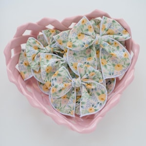 May include: A collection of floral patterned hair bows in a pink heart-shaped dish. The bows feature a white background with yellow, blue, and green floral designs. The dish has a wavy edge and is filled with the bows.