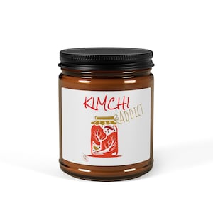 Könnte beinhalten: Ein braunes Glas mit schwarzem Deckel. Das Glas hat ein weißes Etikett mit den Worten "KIMCHI ADDICT" in Rot und Gold. Das Etikett zeigt auch eine rote Illustration von Kimchi in einem Glas.