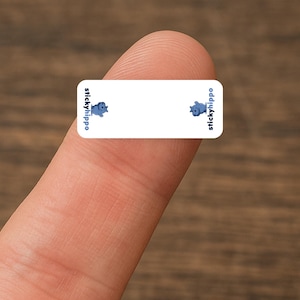May include: A small, rectangular sticker with rounded corners, held by a finger. The sticker is white with blue text and cartoon hippopotamus illustrations. The text "stickyhippo" appears on the left and right sides.