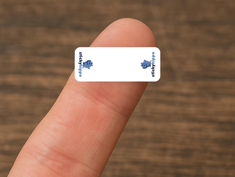May include: A small, rectangular sticker with rounded corners, held by a finger. The sticker is white with blue text and cartoon hippopotamus illustrations. The text "stickyhippo" appears on the left and right sides.