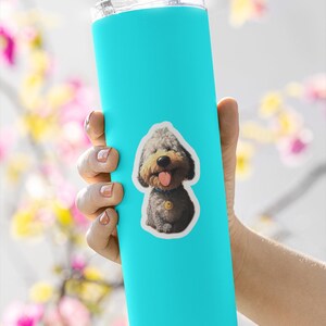 May include: A bright turquoise cylindrical tumbler featuring a sticker of a fluffy gray and tan dog with a blue collar and a yellow tag. The dog has a black nose, a pink tongue sticking out, and a cheerful expression. The tumbler has a clear lid.