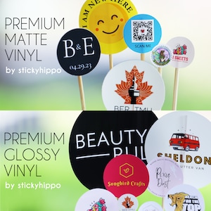 May include: A collection of circular matte and glossy vinyl stickers attached to wooden sticks. The stickers feature various designs including a yellow smiley face, a QR code, a maple leaf, a red van, and text such as B&E 04.29.23, BEAUTY PULSE, and Songbird Crafts.