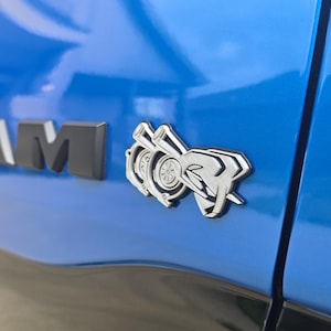 May include: A close-up of a blue vehicle's side panel. The image shows the black and silver "RAM" emblem and a silver emblem with a stylized turbocharger design. The vehicle's paint is a bright, glossy blue.