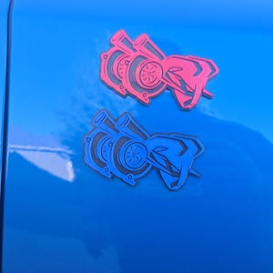 Może przedstawiać: Dwie naklejki z motywem turbosprężarki, jedna różowa i jedna niebieska, są przymocowane do jasnoniebieskiej powierzchni. Naklejki przedstawiają stylizowany wzór turbosprężarki z charakterystycznym obrysem. Tło jest jednolite, błyszcząco niebieskie.