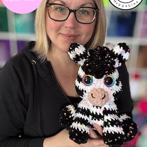 Op de afbeelding: Een vrouw houdt een zwart-wit gebreid zebra knuffeldier vast met regenboogogen. De zebra heeft zwart-witte strepen en kleurrijke confetti-sprinkels door het hele breiwerk. De tekst "Zarah the Zebra Digital Pattern" staat in een roze cirkel in de linkerbovenhoek van de afbeelding. De tekst "Rachel's Handmade Huggables" staat in een zwarte cirkel met een witte ster in de rechterbovenhoek van de afbeelding. De tekst "Digital item not physical item" staat onderaan de afbeelding.