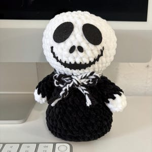 May include: A handmade crochet plush doll featuring a round white head with black felt eyes and a stitched black mouth. The doll wears a black crocheted body with white hands and a black and white braided yarn bow tie around its neck area.