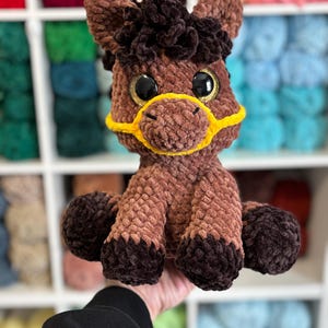 A finished Hollie the Horse plushie variation with rich chocolate and tan tones, golden glittery safety eyes, and yellow halter. Displayed against a wall of colorful yarn for inspiration.