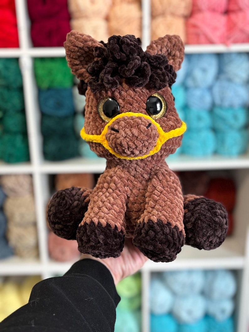 A finished Hollie the Horse plushie variation with rich chocolate and tan tones, golden glittery safety eyes, and yellow halter. Displayed against a wall of colorful yarn for inspiration.
