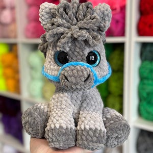 A finished Hollie the Horse plushie variation with soft grey tones, blue glittery safety eyes, and blue halter. Displayed against a wall of colourful yarn for inspiration.