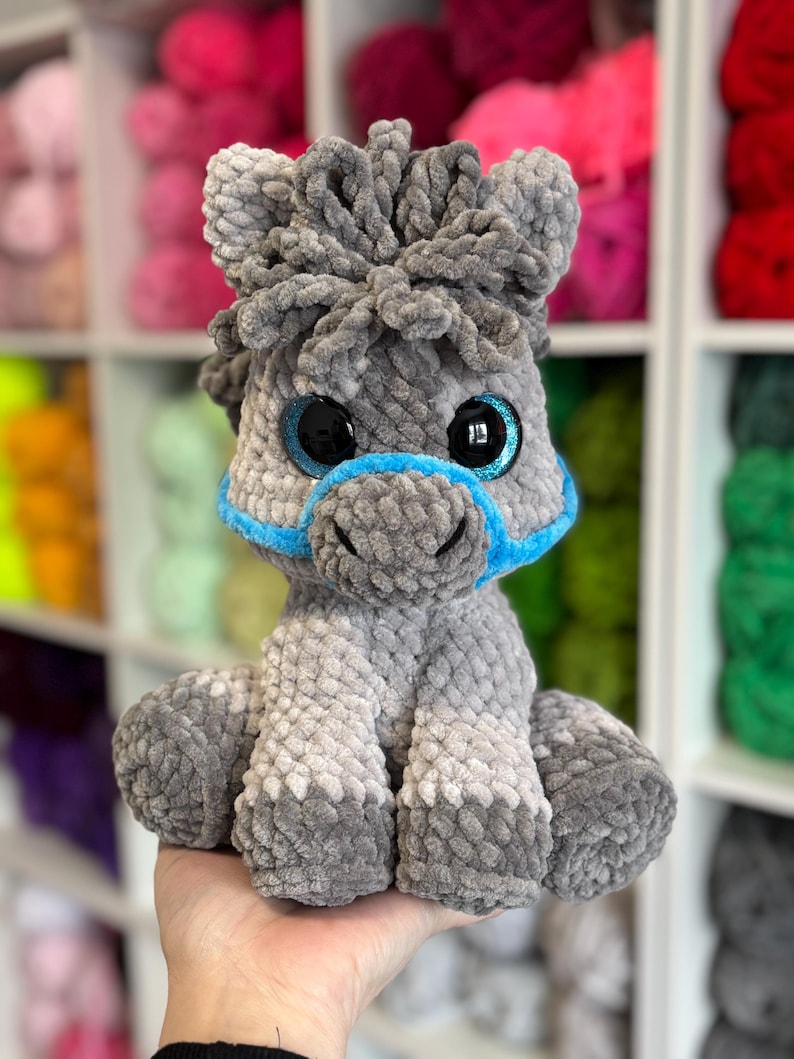 A finished Hollie the Horse plushie variation with soft grey tones, blue glittery safety eyes, and blue halter. Displayed against a wall of colourful yarn for inspiration.