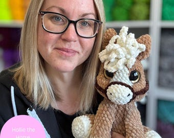 May include: A woman holds a handmade, crocheted horse plushie. The horse is brown with a white mane and muzzle, and has large, expressive eyes. The image includes text that reads "Hollie the Horse Digital Pattern" and "Digital item not physical item."