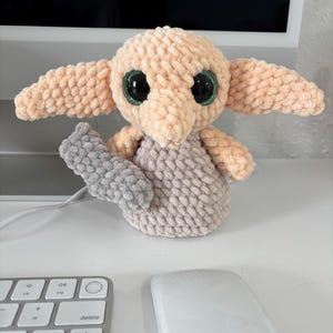 May include: A plush toy made of soft, peach-colored yarn with large, dark, glossy eyes and wide, pointed ears. The figure has a light gray, textured body and holds a small, rectangular, gray, textured object in its right hand.