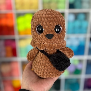 May include: A brown crocheted plush toy with a rounded head and body, featuring large black eyes with glittery pink rims and a small black nose. The plush figure wears a small black crocheted satchel bag across its chest, crafted from soft yarn.