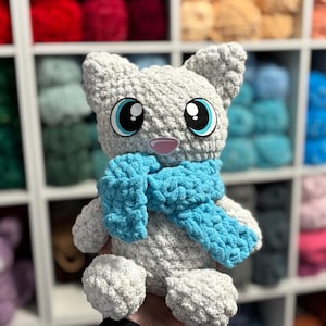 Może przedstawiać: Ręcznie robiona szara pluszowa zabawka w kształcie kota z dużymi niebieskimi oczami i jasnoniebieskim szalikiem. Kot ma różowy nos i jest wykonany z miękkiej, teksturowanej przędzy. Pluszak to urocza, przytulna zabawka.