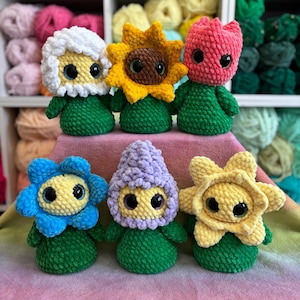 May include: Six handmade crochet dolls, each with a unique flower or plant-themed design. The dolls feature green bodies, large eyes, and various flower or plant-shaped heads in colors like yellow, blue, pink, and white. The background includes shelves of colorful yarn.
