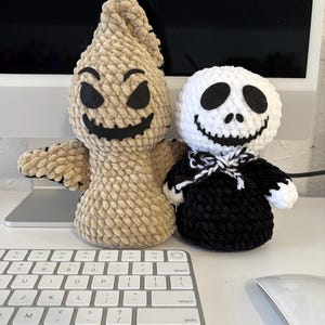 May include: Two handmade crochet plush toys. One is a tan, sack-like figure with a pointed top, black felt eyes, and a stitched black mouth. The other is a round-headed figure with a white face, black felt eyes, a stitched mouth, and a black body.