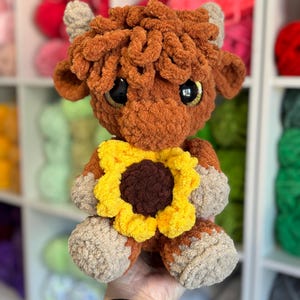 Op de afbeelding: Een handgemaakte pluche Highland-koe met bruine vacht, bruine hoeven en een gele en bruine zonnebloemkraag. De koe heeft grote, expressieve ogen en kleine, grijze hoorns. Het pluche speelgoed is zacht en knuffelig.