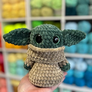 May include: A handmade plush doll in the likeness of a character with large eyes and pointed ears. The doll is primarily green and tan, with large black eyes. The plushie is made of a soft, textured material, and is a popular character from a science fiction franchise.