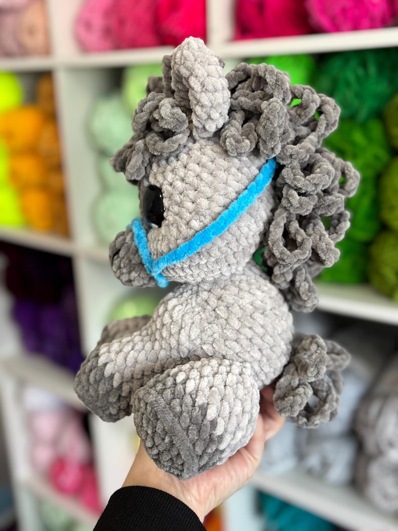 Side view of a gray Hollie the Horse plushie with a curly mane and tail, wearing a blue halter. Displays the finished plushie’s soft texture and charming shape.