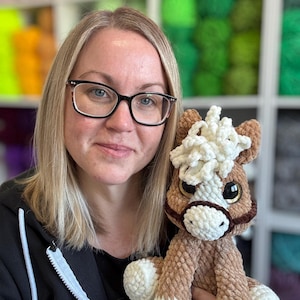 Designer holding Hollie the Horse plushie with cream mane and white hooves, smiling in front of bright yarn shelves. Helps reinforce that this is a handmade digital pattern product.
