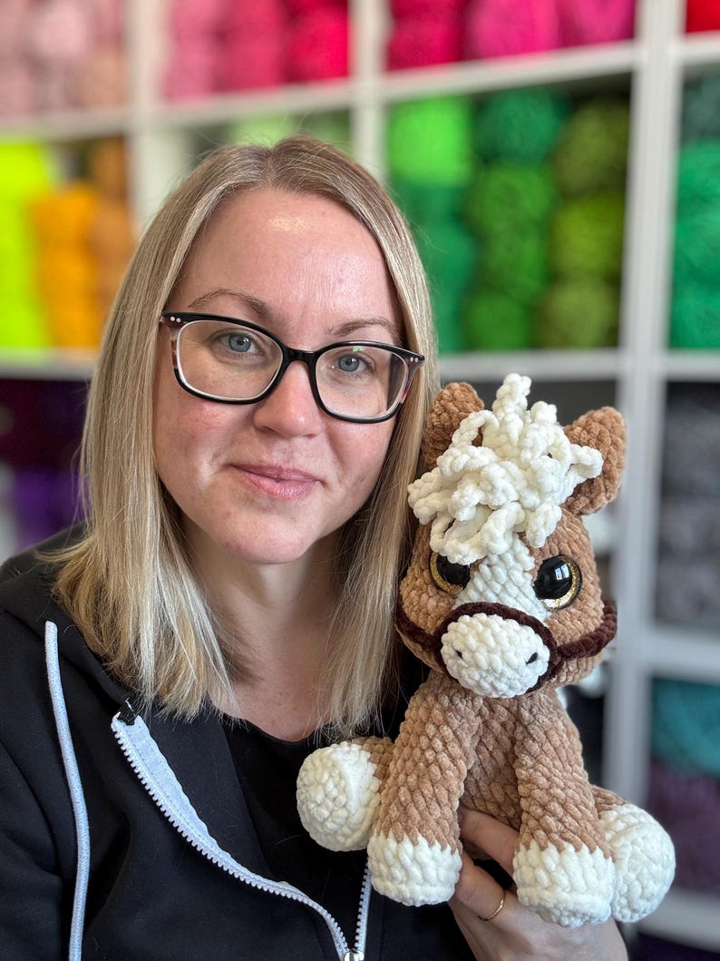 Designer holding Hollie the Horse plushie with cream mane and white hooves, smiling in front of bright yarn shelves. Helps reinforce that this is a handmade digital pattern product.