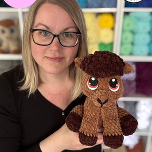 May include: A woman holds a brown crocheted camel plush toy with large red eyes. The toy is sitting on her hand. The background is a wall of colorful yarn balls. The text "Cody the Camel Digital Pattern" is in a pink circle at the top left of the image. The text "Rachel's Handmade Huggables" is in a circle with a black "R" in the top right of the image.