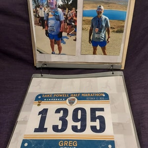 May include: A blue and white race bib with the text "LAKE POWELL HALF MARATHON" and "OCTOBER 12, 2019". The number "1395" is printed in large blue font. The text "GREG #LPHALF" is printed below the number.