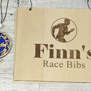 May include: A wooden sign with the text "Finn's Race Bibs" and a silhouette of a runner. The sign is hanging from two hooks. A wooden medal with a blue center and the text "Finnegan" is hanging from a string.