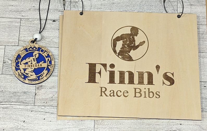 May include: A wooden sign with the text "Finn's Race Bibs" and a silhouette of a runner. The sign is hanging from two hooks. A wooden medal with a blue center and the text "Finnegan" is hanging from a string.