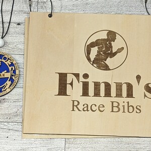 Pu&ograve; includere: Un cartello di legno con il testo "Finn's Race Bibs" e una silhouette di un corridore. Il cartello &egrave; appeso a due piccoli ganci di metallo. Una medaglia di legno con un centro blu e il testo "Finnegan" &egrave; appesa a una corda.