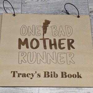 Pu&ograve; includere: Una copertina di libro in legno con il testo "One Bad Mother Runner" e "Tracy's Bib Book" in lettere marroni. La copertina ha due cinturini neri con anelli metallici.