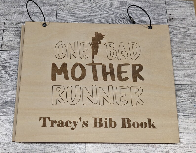 Pu&ograve; includere: Una copertina di libro in legno con il testo "One Bad Mother Runner" e "Tracy's Bib Book" in lettere marroni. La copertina ha due cinturini neri con anelli metallici.