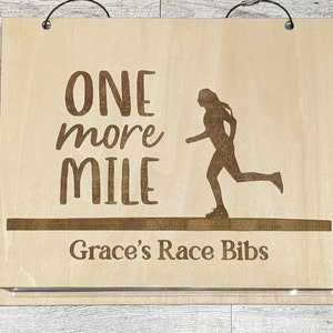Puede incluir: Un letrero de madera con el texto "One more mile" y una silueta de un corredor. El letrero también tiene el texto "Grace's Race Bibs".