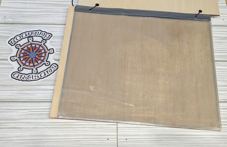 Pu&ograve; includere: Una lavagna rettangolare, vuota, di colore marrone chiaro, incorniciata con un coperchio in plastica trasparente. La lavagna &egrave; montata su una superficie in legno chiaro.
