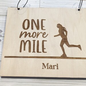 Pu&ograve; includere: Un cartello di legno con il testo "ONE more MILE" e una silhouette di un corridore. Il nome "Mari" &egrave; scritto sotto il testo.