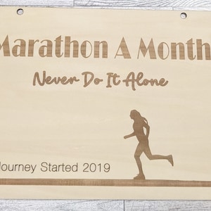 Pu&ograve; includere: Un cartello di legno con il testo "Marathon A Month Never Do It Alone" e una silhouette di un corridore. Il cartello include anche il testo "Journey Started 2019".