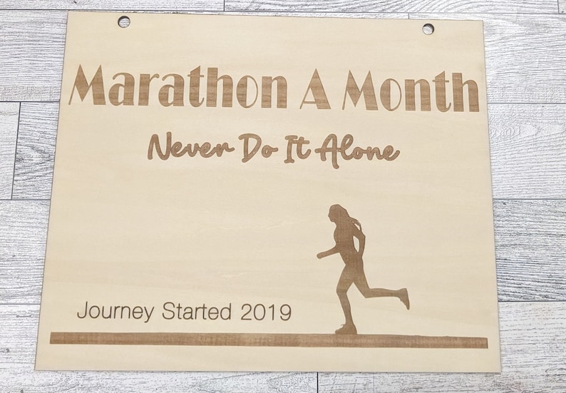 Pu&ograve; includere: Un cartello di legno con il testo "Marathon A Month Never Do It Alone" e una silhouette di un corridore. Il cartello include anche il testo "Journey Started 2019".