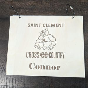 May include: A wooden plaque with the text "SAINT CLEMENT CROSS COUNTRY Connor" and a cartoon mascot of a tiger wearing a wrestling singlet.