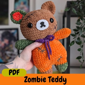 Könnte beinhalten: Ein gehäkelter Zombie-Teddybär mit braunem Fell, orangefarbener und grüner Kleidung und einem lila Band. Der Bär hat angenähte Augen und eine weiße Nase. Das Bild enthält den Text "PDF Zombie Teddy".