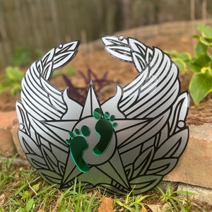 May include: A silver metal ornament shaped like a star with wings and a green footprint design in the center.
