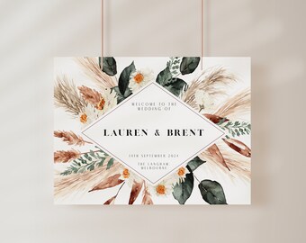 May include: A wedding welcome sign with a white background and a diamond-shaped frame. The frame features the text "Welcome to the wedding of Lauren & Brent" and the date "14th September 2021". The background is decorated with watercolor illustrations of pampas grass, eucalyptus leaves, and white flowers.