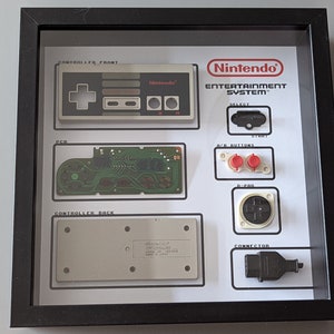 May include: A black framed display featuring a disassembled Nintendo Entertainment System controller. Components include the front and back casing, a green circuit board, red A/B buttons, a black D-pad, a start/select button, and a black connector.