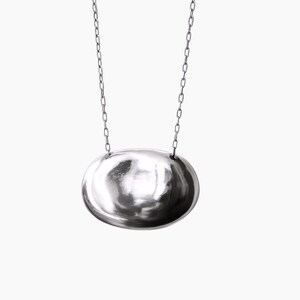 May include: A silver necklace with an oval pendant hanging from a fine chain. The pendant has a polished, reflective surface, creating a bright, metallic appearance. The chain is also silver, matching the pendant's design. The necklace is set against a plain white background.