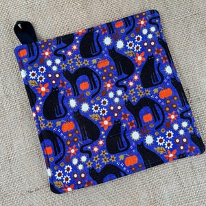 May include: A square fabric potholder featuring a vibrant blue background patterned with black cat silhouettes, orange pumpkins, and small white and yellow flowers. The potholder includes a black fabric loop attached to one corner for hanging.