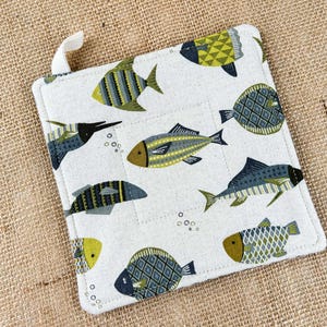 May include: Square white pot holder with a loop, featuring a repeating pattern of stylized fish in shades of green, gray, and yellow. The fish designs vary in shape and pattern, creating a whimsical, aquatic theme.