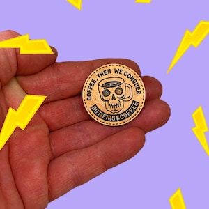 以下が含まれることがあります： 「COFFEE, THEN WE CONQUER BUT FIRST COFFEE」の文字が、コーヒーカップとドクロのデザインを囲むように刻まれた、丸い銅色のコイン。紫色の背景に黄色の稲妻模様が描かれ、手でコインが持たれています。