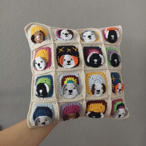 Op de afbeelding: Een vierkant, crèmekleurig gehaakt kussen met een raster van zestien vierkanten, elk met een ander honden gezicht. De honden hebben verschillende kleuren en patronen, waaronder bruin, zwart en wit. Het kussen wordt door een hand vastgehouden.