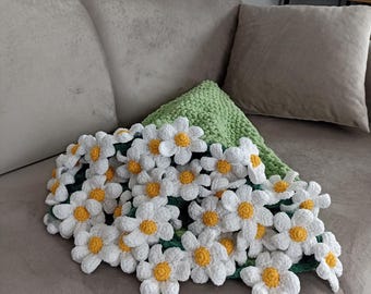 May include: A crocheted daisy blanket with white petals and yellow centers, resting on a green crocheted pillow. The blanket is adorned with numerous daisy flowers. The items are placed on a gray sofa with a matching pillow.