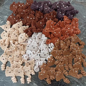 May include: A collection of handmade crochet figures in various shades of brown, beige, and white. Each figure has a smiling face and a star-like pattern. The figures are arranged in a pile, showcasing the variety of colors and designs.